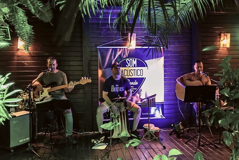 Alexandre no contrabaixo, Robson no cajón e Alan Peter no violão em um deck de madeira cercado de plantas