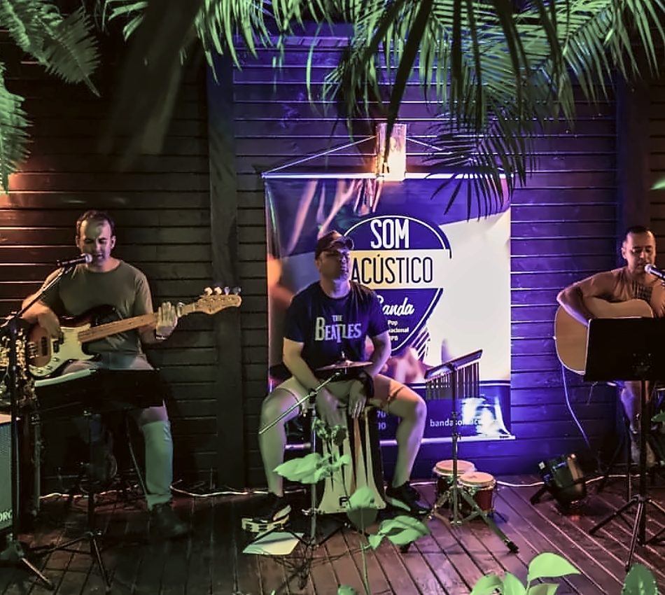 Alexandre no contrabaixo, Robson no cajón e Alan Peter no violão tocando em um deck de madeira, com o banner da banda ao fundo